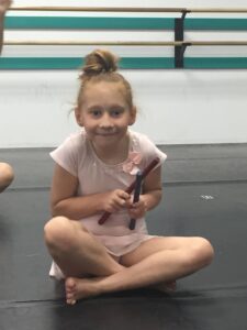 Dance student wearing a pink dance uniform is sitting in a dance studio holding a red and a blue stick.